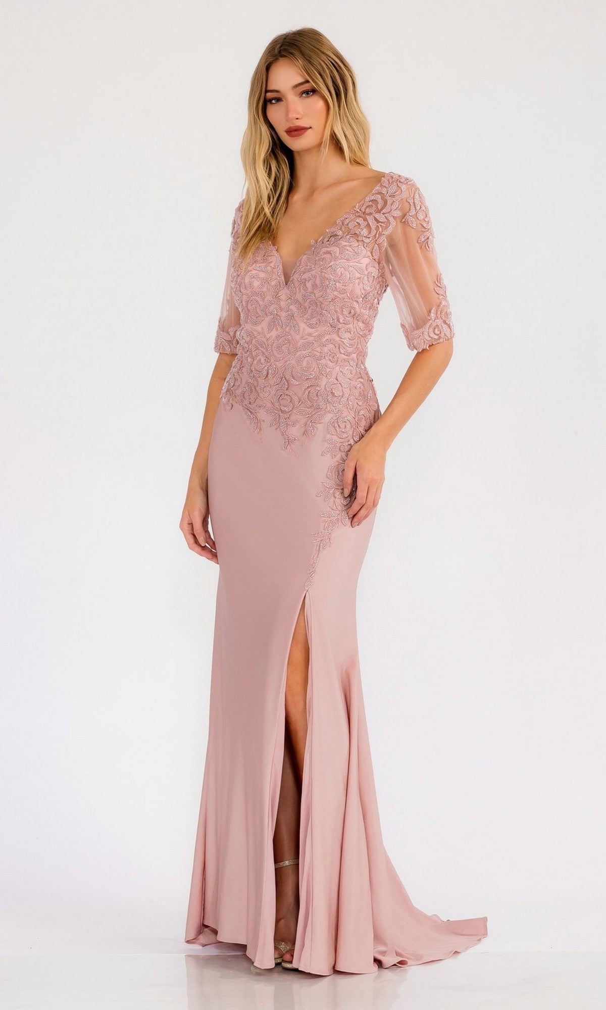 A woman wearing the Juliet JTM15L, a rose-colored, short sleeve long prom dress featuring beaded embroidery, lace details, a deep V-neckline, and a front slit, stands against a plain white background.